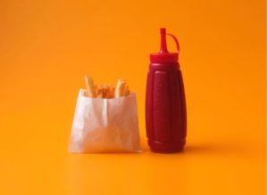 A plastic bottle of ketchup besides a bag of fries on an orange background.